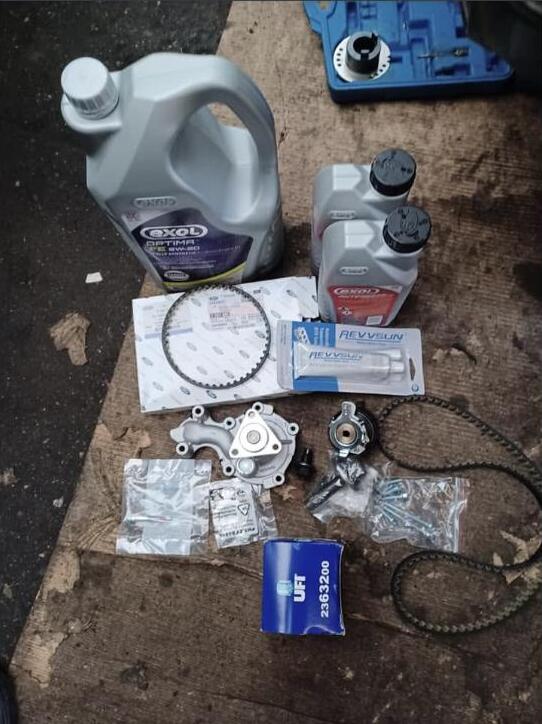 The parts for a wet timing belt job laid out on the workshop floor: a can of Exol Optima 5W-20 oil, two smaller bottles, a new toothed belt, a water pump, an oil filter, a tube of sealant, bagged bolts and a blue case of engine timing tools.
