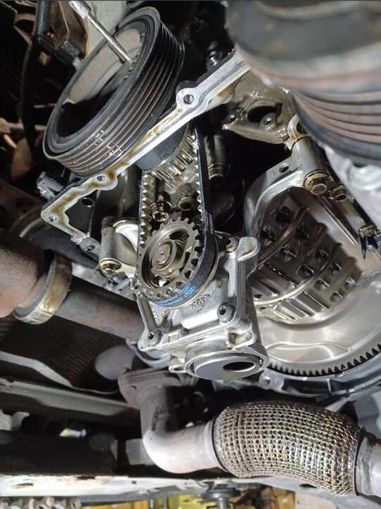 The lower end of the engine seen from underneath, showing the crankshaft sprocket, the new belt running down to it, the oil pump housing and the auxiliary belt pulley above.