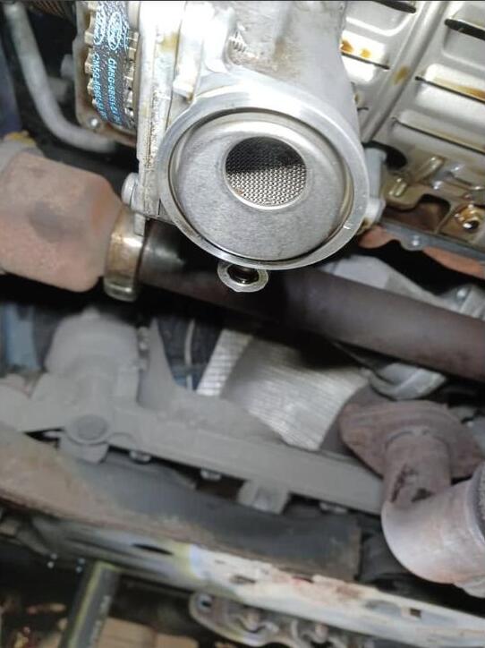The oil pickup pipe removed and held up to the camera, its fine mesh strainer clean and clear, with the exhaust and subframe behind it.