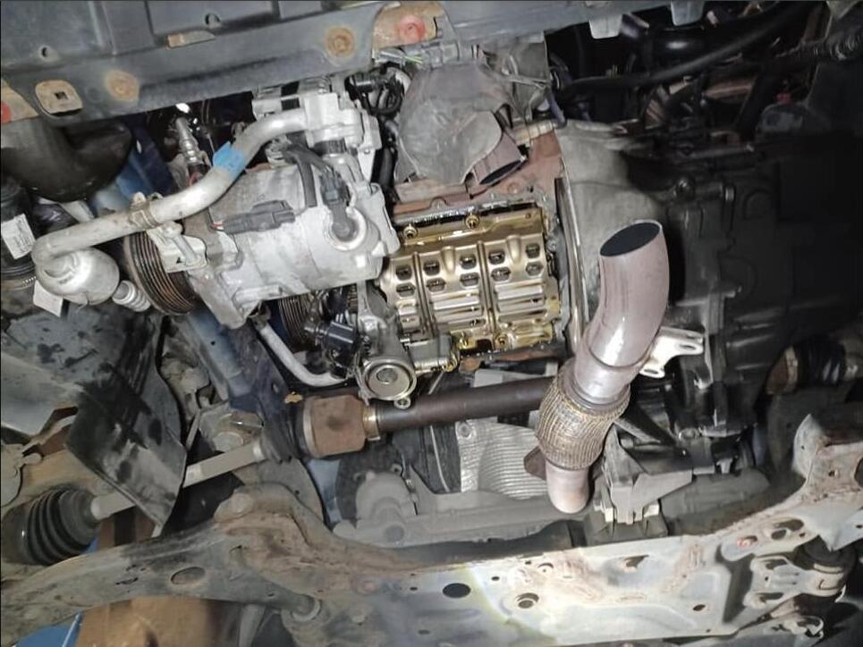 Wide view of the underside of the car with the sump removed, showing the exposed oil pump and baffle, the driveshaft, the exhaust downpipe and the subframe.